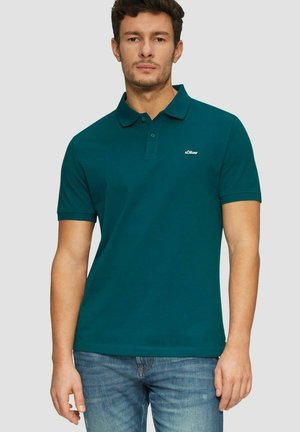 Junger Mann trägt ein türkisfarbenes Poloshirt mit "s.Oliver"-Logo auf der Brust und blaue Jeans, steht vor einem schlichten hellgrauen Hintergrund.