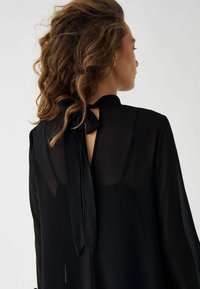 Črna bluzica z visokim ovratnikom in vezanjem na hrbtu. Prosojna tkanina z tekočim dizajnom. Dolgi rokavi, minimalistični slog.
