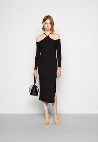Vestido preto com decote à mostra, mangas longas, corte justo, até ao meio da perna, com fenda lateral. Combinado com sandálias amarelas de tiras e uma pequena mala preta.