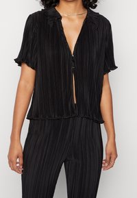 Zwarte blouse met korte mouwen, een geplooid ontwerp, een kraag en een knoopsluiting aan de voorkant. Lichte en flowy textuur.