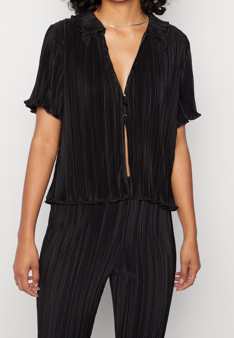 Černá blůza s krátkým rukávem, plisovaným designem, límečkem a zapínáním na knoflíky vpředu. Lehká a splývavá textura.