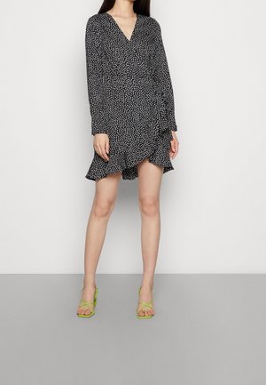 Woman wearing a black wrap dress with white polka dots and ruffled hem, paired with lime green strappy high-heeled sandals.