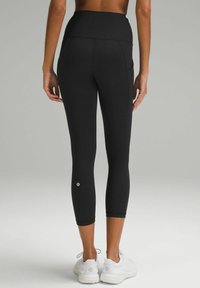 Leggings neri a vita alta con una texture liscia, taglio a lunghezza ginocchio e logo accentuato. Abbinati a scarpe da ginnastica bianche.
