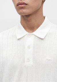 Gros plan d'une personne portant un polo blanc en maille avec des rayures verticales et trois boutons sur la patte de boutonnage.