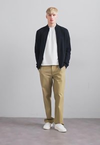 Tânăr cu părul blond, îmbrăcat cu o jachetă neagră, cămașă albă, pantaloni bej și adidași albi, stând în fața unui fundal gri uniform.