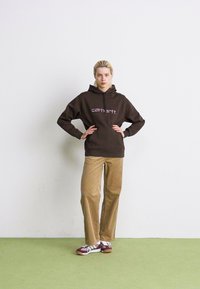 Vrouw staat met handen op de heupen, gekleed in een donkerbruine Carhartt hoodie, beige corduroy broek en bordeauxrode sneakers op een groene vloer.