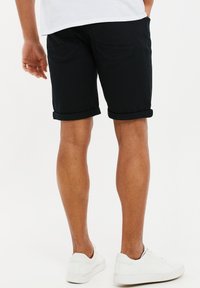 Homme portant un short noir retroussé, des baskets blanches et une chemise blanche, se tenant contre un fond blanc uni.