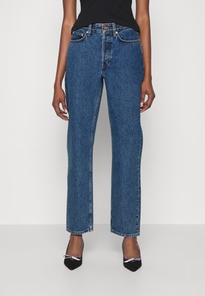Person trägt gerade geschnittene blaue Denim-Jeans, schwarze spitze High Heels mit Schleifen und ein schwarzes ärmelloses Top, steht vor einem einfarbigen Hintergrund.