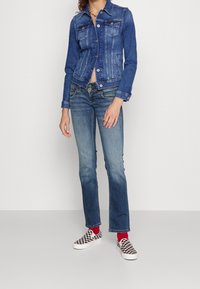 Džínová bunda s přiléhavým střihem, světle modrým efektem a knoflíkovými detaily. Kombinována s rozšířenými modrými džínami a černobílými nazouvacími botami se šachovnicovým vzorem.