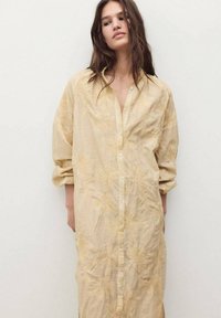 Donna che indossa un lungo e ampio vestito beige con bottoni e ricami floreali, in piedi contro un muro leggero e uniforme, con le braccia rilassate lungo i fianchi.