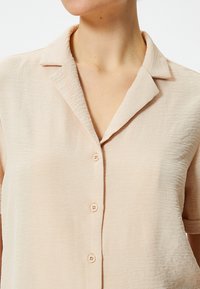 Beige korte mouwen shirt van gekreukt stof; heeft een klassieke kraag en vijf lichte knopen langs de voorkant voor sluiting.