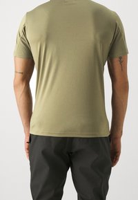 Olivově zelené tričko s krátkým rukávem a plochým švem, vyrobené z hladké látky. Viditelné tetování na horní části paže. Nosené s tmavými kalhotami.