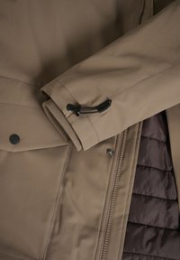 Giacca esterna marrone con tessuto testurizzato, dotata di chiusura con zip, dettagli con bottoni e foderatura interna trapuntata di una tonalità più scura.