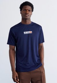 Laivyno mėlynos spalvos trumpomis rankovėmis t-shirt, pagaminta iš lengvos medžiagos, su priekiniu baltu ir oranžiniu logotipu. Lygus paviršius, su apvaliu kaklu.