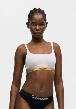 Donna con i capelli intrecciati che indossa un reggiseno bianco Calvin Klein con banda arcobaleno e biancheria intima nera Calvin Klein su uno sfondo semplice.