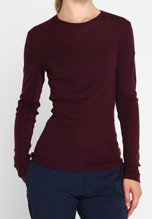 Kobieta ubrana w dopasowaną, długą bluzkę z okrągłym dekoltem w ciemnym bordowym kolorze oraz granatowe spodnie, stojąca z jedną ręką w kieszeni.