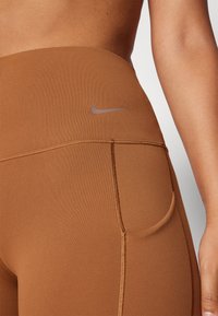 Smeđe tajice visokog struka s glatkom teksturom, koje sadrže skriveni džep i reflektirajući Nike logo na pojasu.