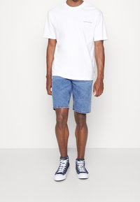 Camiseta blanca de manga corta con un pequeño gráfico, combinada con pantalones cortos de mezclilla azules y zapatillas altas marino con suelas blancas.