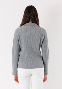 Maglione grigio a costine con colletto alto, corpo aderente e maniche lunghe, indossato sopra pantaloni bianchi. Tessuto testurizzato con linee verticali.