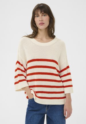 Femme portant un pull en tricot crème à rayures horizontales rouges et un jean bleu, debout avec une main dans la poche devant un fond uni.
