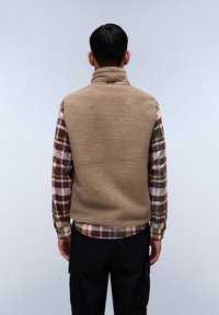Beige fleece vest with high collar, textured surface, and no hood, layered over a plaid shirt in earthy tones. Black cargo pants visible below.