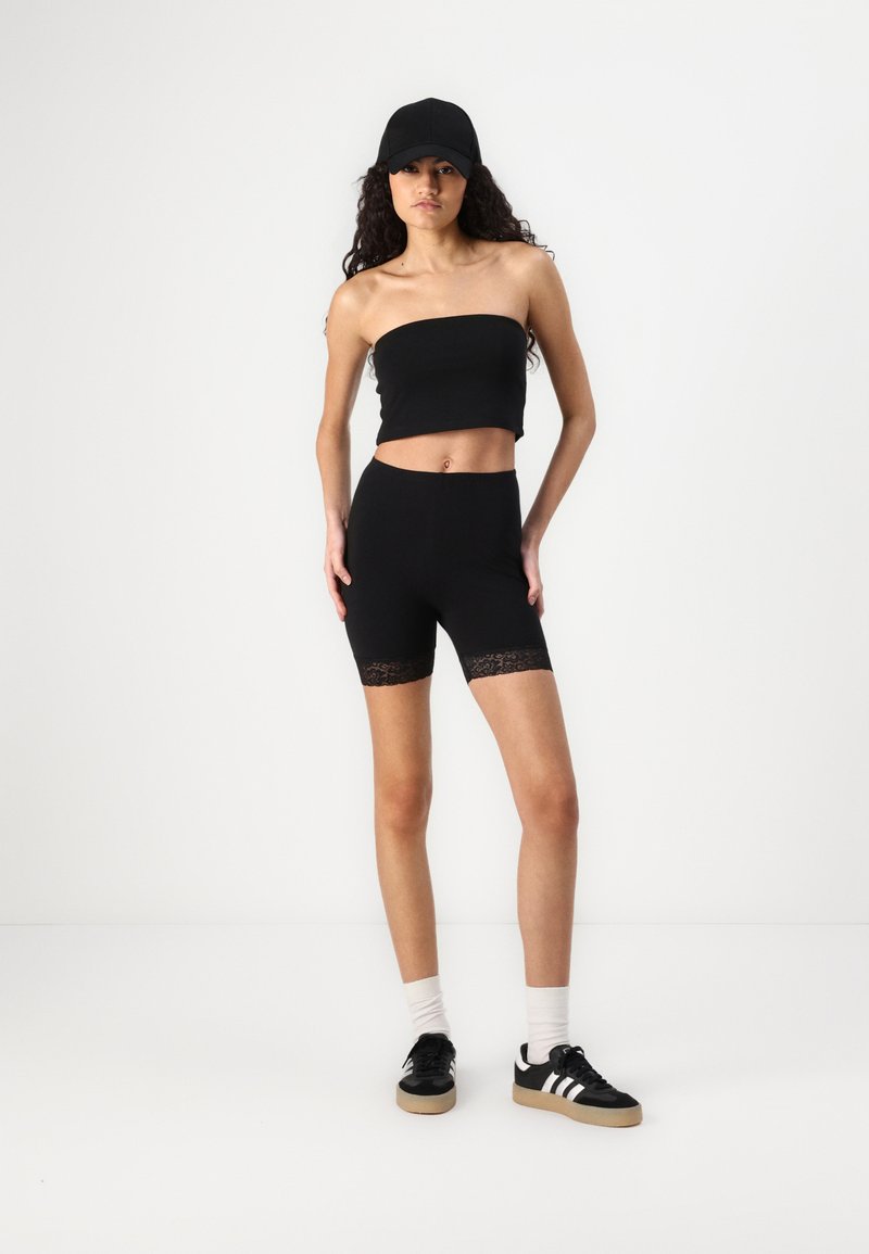Black strapless crop top and high-waisted cycling shorts with lace trim. Model wears black cap and sneakers with white stripes.