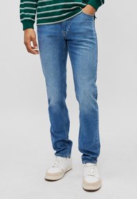 Jeans de denim azul claro com um corte slim, apresentando cinco bolsos e costuras discretas. Combinados com tênis brancos, exibindo um visual descontraído.