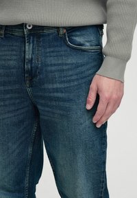 Jeans iz temno modrega denima z rahlo zbledelim pranjem, ki vključujejo pet žepov, vidno šivanje in sproščen kroj. Nosijo se z svetlo sivo puloverjem.