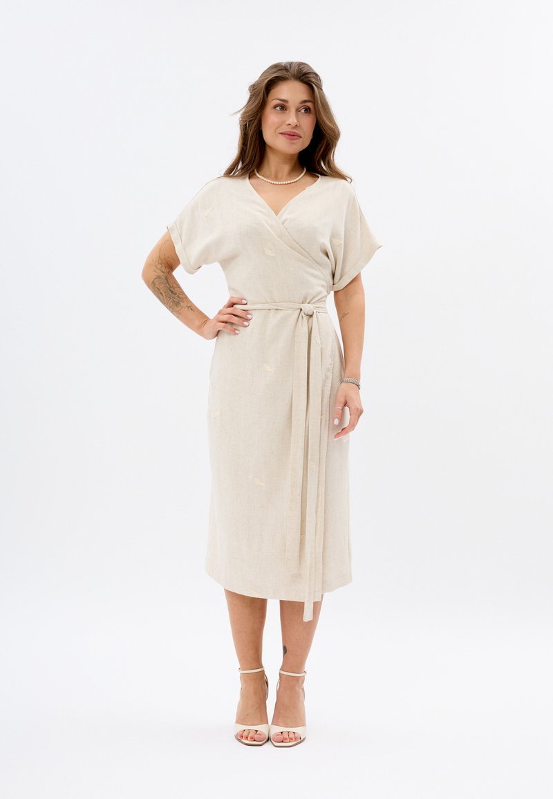 Hellbeige, wickelartiges Kleid aus Leinen, mit kurzen Ärmeln, einem Bindegürtel und dezenten Blättermustern. Getragen mit Riemchen-Pumps.