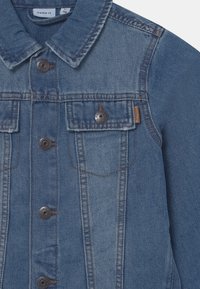 Jeansjacke in Mittelblau mit Kragen, vorderem Knopfverschluss und zwei Brusttaschen mit dunklen Knöpfen und kontrastierenden Nähten.