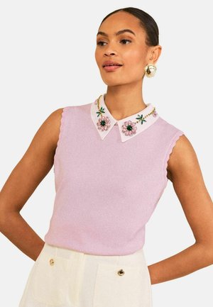 Femme portant un haut sans manches rose clair avec un col blanc orné de détails floraux et de pierres précieuses, associé à un pantalon blanc taille haute.