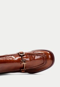 Zapato de charol marrón con un diseño elegante, que presenta acentos de hardware dorado y costuras detalladas a lo largo de los bordes.