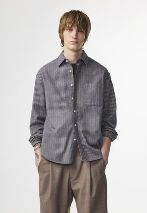 Checkered button-up shirt in blue and beige tones, featuring a chest pocket and rolled sleeves, paired with light brown trousers.