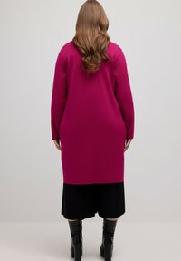 Langer magentafarbener Pullover mit geradem Schnitt, mit langen Ärmeln und glatter Textur, getragen über einem schwarzen Plisseerock und Stiefeletten mit Absatz.