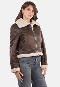 Chaqueta de cuero marrón recortada con un cuello de borreguito falso de color crema, que cuenta con un cierre frontal, bolsillos laterales y detalles de costura en contraste.