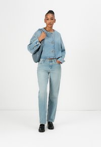 Lichtblauwe denim cropped jas met messing knopen, gecombineerd met lichtblauwe straight-leg jeans en zwarte instappers. Houdt een donkerblauwe tas vast.