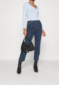 Femme portant un cardigan bleu clair à boutons, un jean floral bleu foncé, des bottes noires à la cheville, tenant un sac à main noir sur fond blanc.