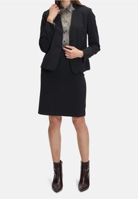 Svart skräddarsydd kavaj över en grå skjorta med knappar, kombinerad med en svart blyertskjol och mörkbruna ankelstövlar. Slät tyg, figursydd design.