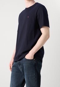 Camiseta de algodón azul marino, de manga corta, con cuello redondo y pequeño detalle de logotipo en el pecho. Combinada con unos jeans de denim oscuro.