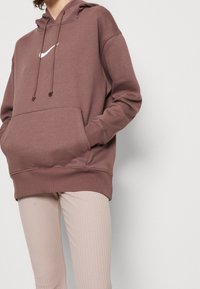 Felpa con cappuccio marrone con logo Nike bianco, dotata di tasca frontale e polsini a costine. Abbinata a leggings chiari a costine.