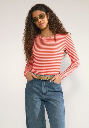 Vrouw met lang krullend haar die groene zonnebril draagt, rood-wit gestreept shirt met lange mouwen, blauwe spijkerbroek en een beige riem met handen in de zakken.