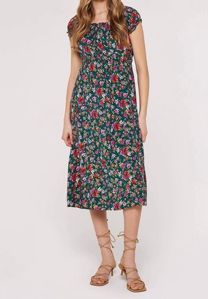 Kvinde iført en grøn midikjole med et flerfarvet blomsterprint og brune sandaler med stropper og høje hæle.