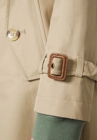 Beige trenchcoatärm med en brunt läderspänne och gyllene knapp. Manschetten har en grön stickad foder för extra textur.