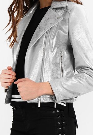 Personne portant une veste de motard argentée métallique par-dessus un top noir à rayures blanches, les mains tenant les bords de la veste, associée à un pantalon noir.