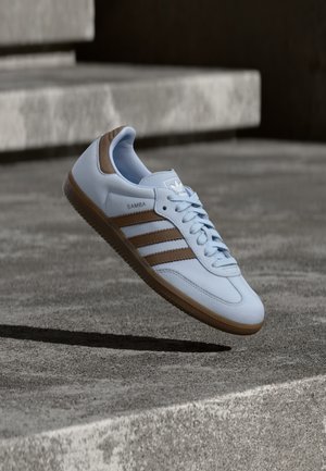 Light blue sneaker with brown suede accents, featuring three lateral stripes, flat laces, and a textured gum sole. Brand logo on tongue.