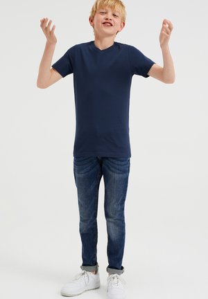 Mörkblå V-ringad t-shirt i bomull, kombinerad med slim-fit blå jeans och vita sneakers, där individen poserar med uppsträckta armar.