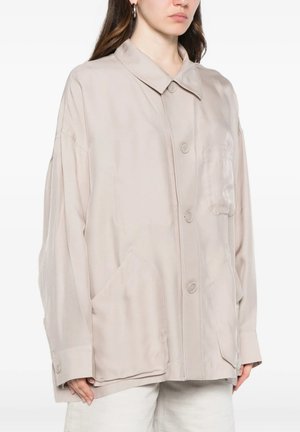 Donna con indosso una giacca beige oversize con bottoni, grandi tasche frontali e colletto, in piedi davanti a uno sfondo semplice.