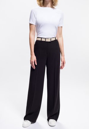 Donna che indossa una maglietta bianca a maniche corte, pantaloni neri a gamba larga con una cintura beige e sneakers bianche, in piedi davanti a uno sfondo neutro.