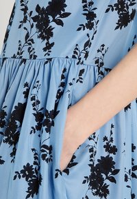 Gros plan d'une main reposant dans la poche d'une robe bleue avec des motifs floraux noirs et un détail froncé à la taille.