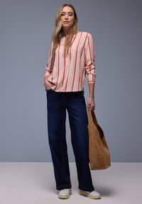 Femme aux longs cheveux blonds portant un chemisier rayé corail et blanc, un jean large bleu foncé, des chaussures blanches, tenant un sac fourre-tout en daim marron.